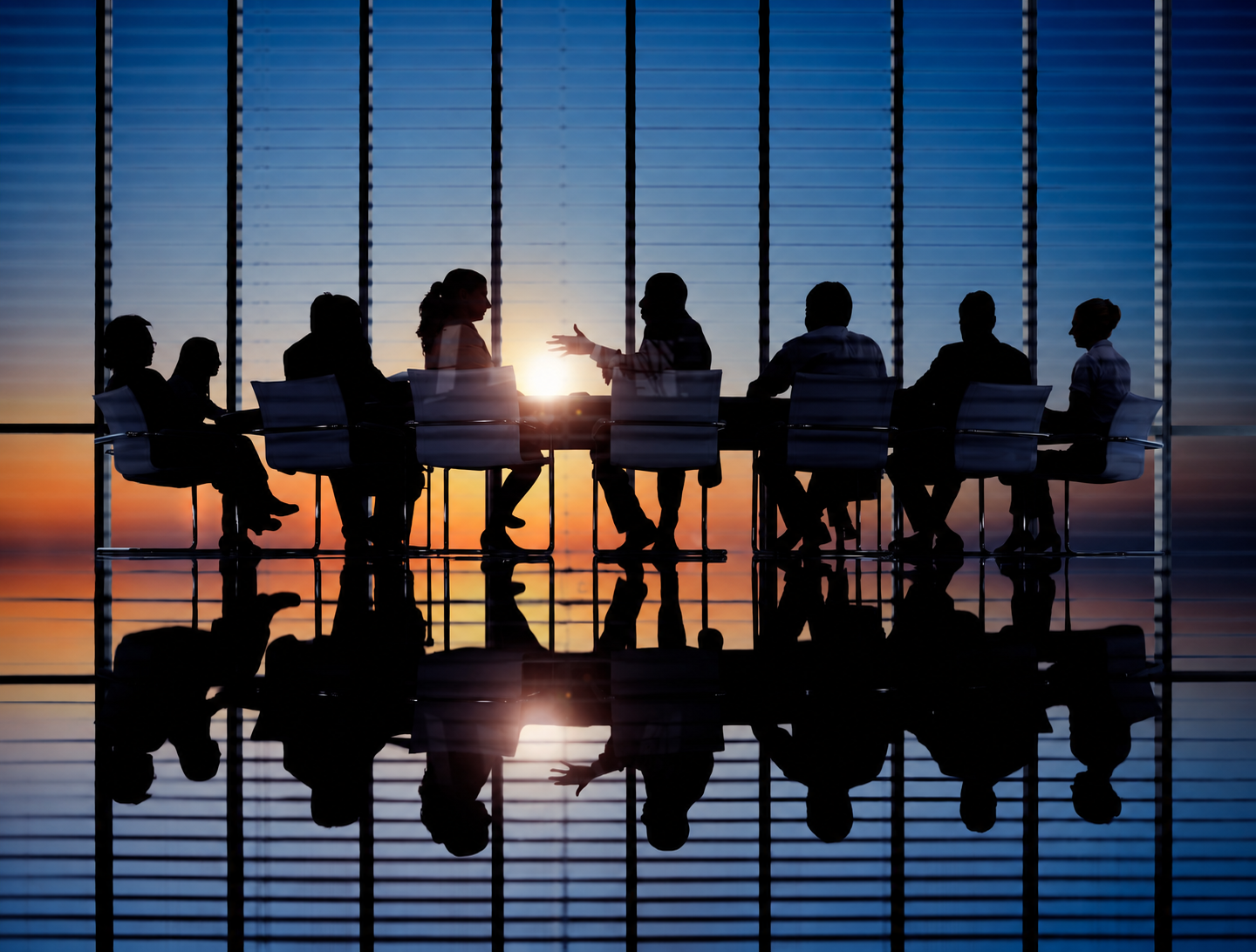 Business leaders in discussion around a conference table, silhouetted against sunset windows.