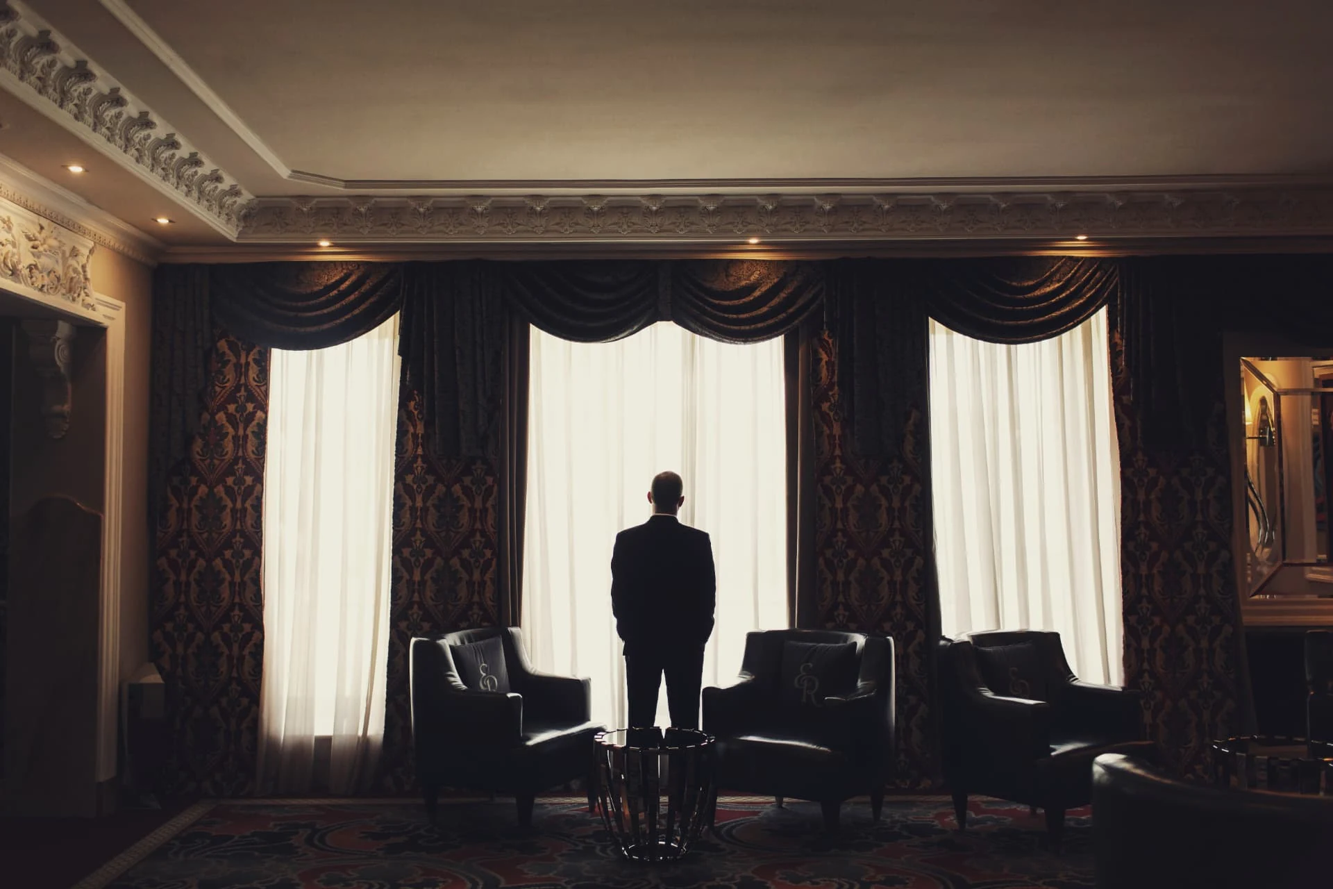 A man standing before tall windows in a warmly lit, elegant room.